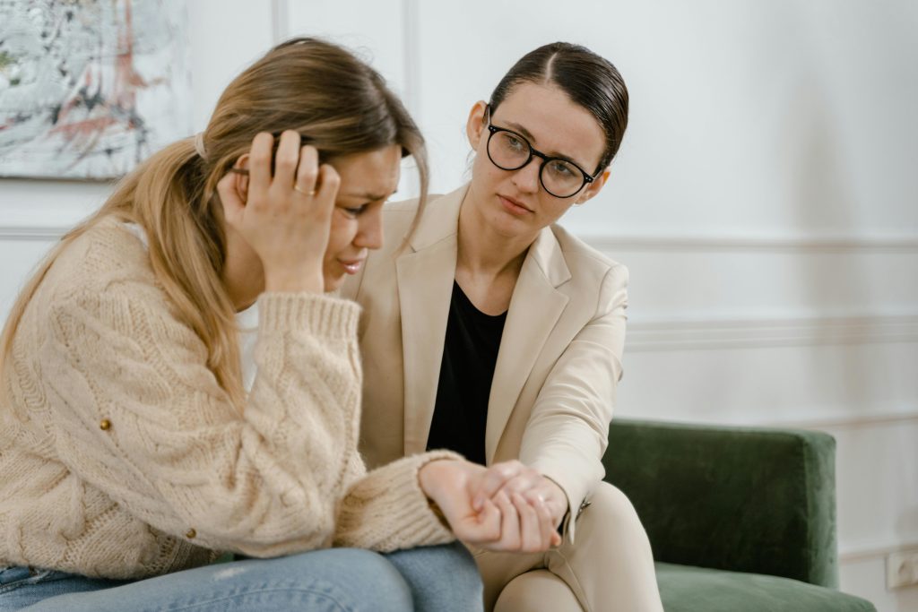 Two women sitting on couch, one upset, one holding the other's hand